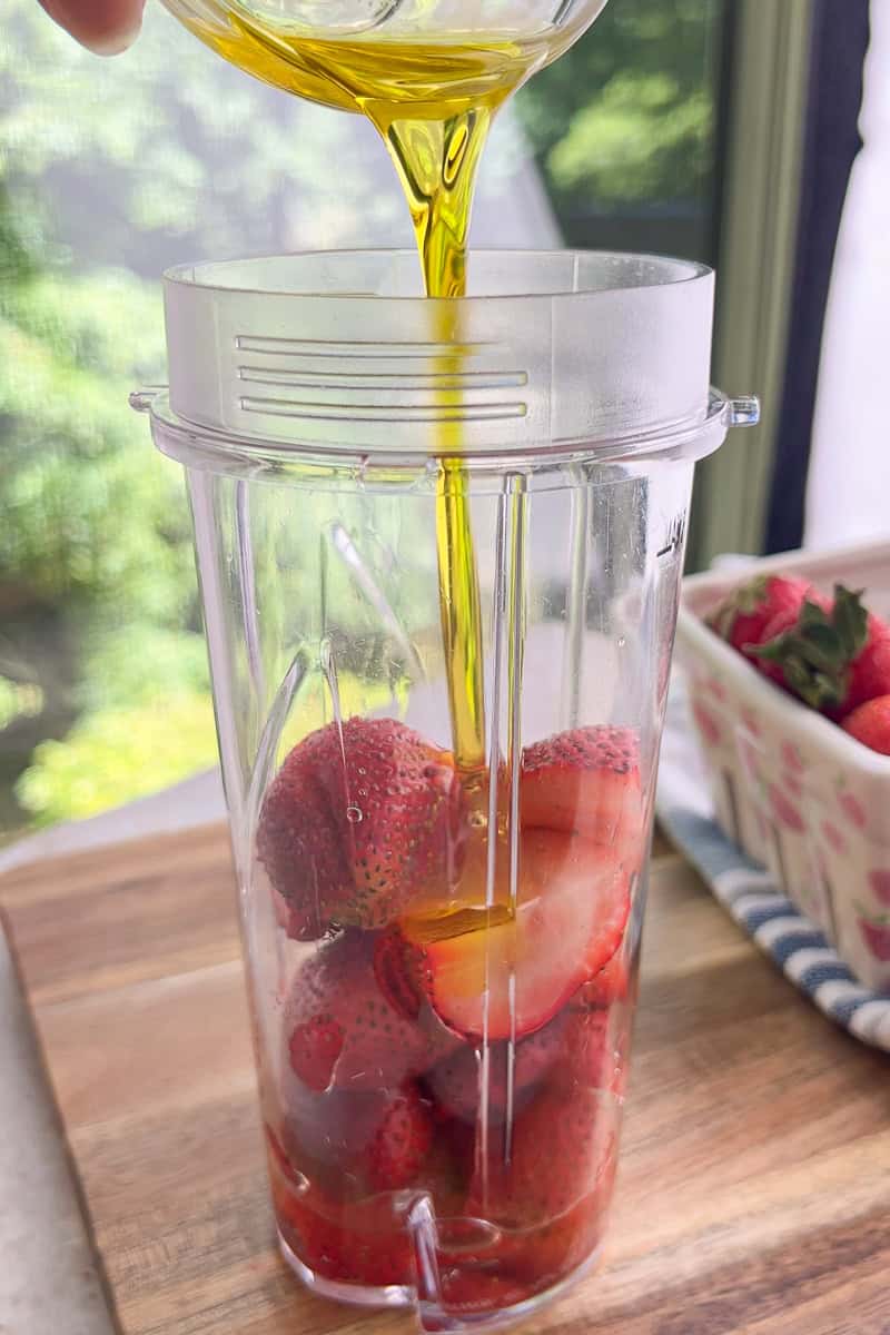Olive oil is poured into a blender with fresh strawberries for homemade strawberry poppyseed dressing.