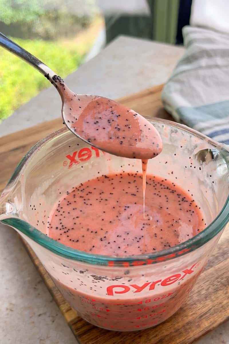 A spoon lifts creamy strawberry poppyseed dressing from a glass measuring cup.