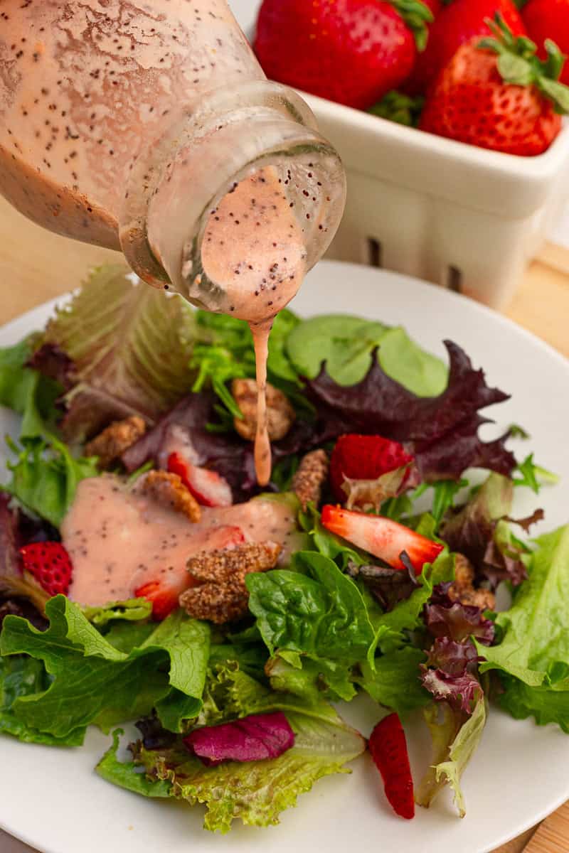 Strawberry poppyseed dressing is poured over a green salad with fresh strawberries and candied pecans.