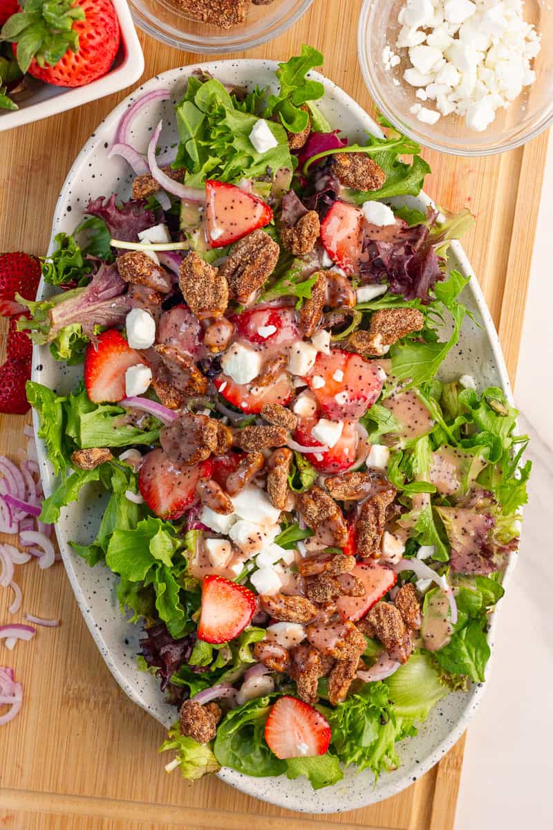 A strawberry salad with goat cheese, cinnamon pecans, red onion, and dressing is served on a platter.