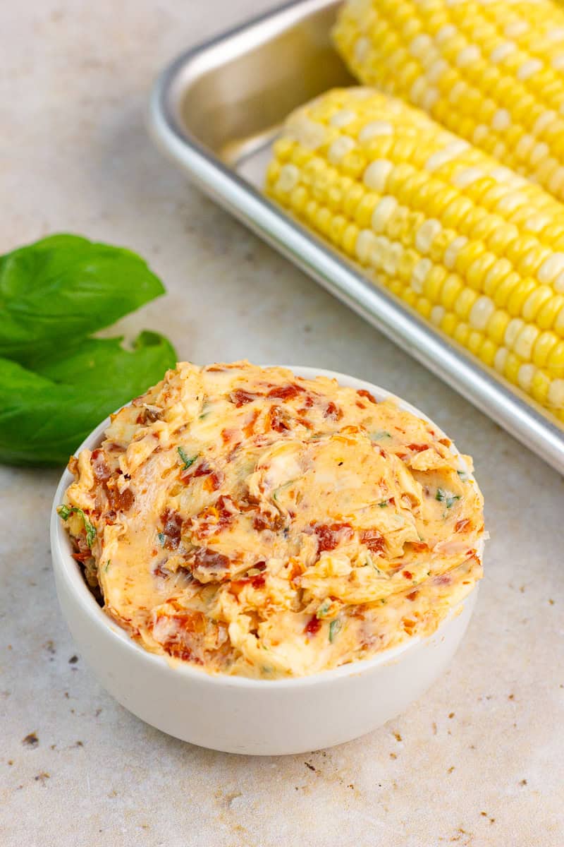 Tomato basil butter is served in a small white bowl with fresh basil and corn on the cob in the background.
