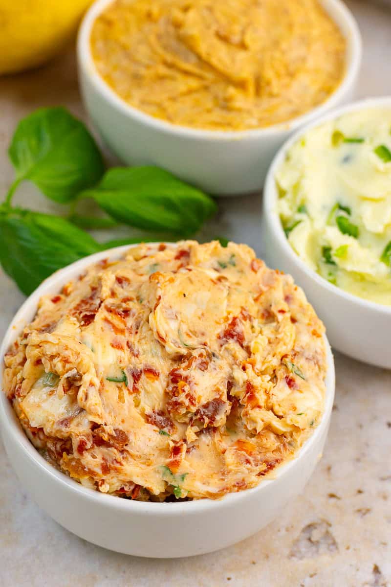 Tomato basil butter is shown in a white bowl with other flavored butters and fresh basil nearby.