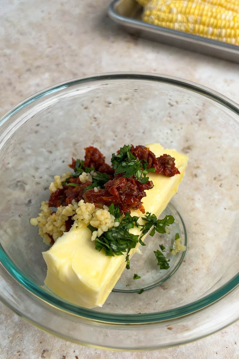 Softened butter, sun-dried tomatoes, garlic, and fresh basil are added to a glass mixing bowl.
