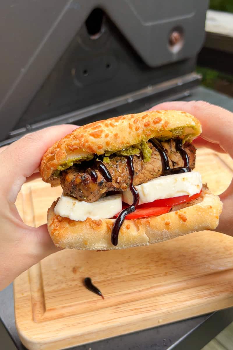Hands hold a finished chicken Caprese sandwich filled with pesto, balsamic chicken, mozzarella, tomato, and balsamic glaze.
