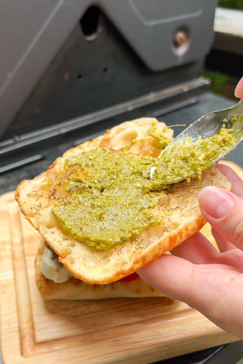 Pesto is spread onto toasted ciabatta as the chicken Caprese sandwich is assembled.