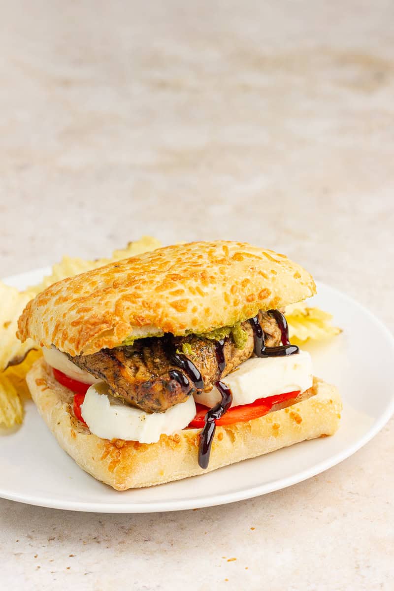 A chicken Caprese sandwich with pesto, mozzarella, tomato, and balsamic glaze is served on a white plate with ridged potato chips.