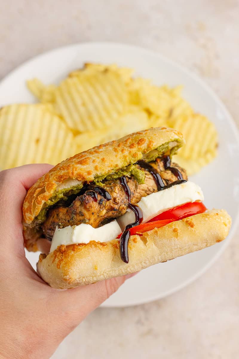 A hand holds a chicken Caprese sandwich filled with grilled balsamic chicken, pesto, mozzarella, tomato, and balsamic glaze.