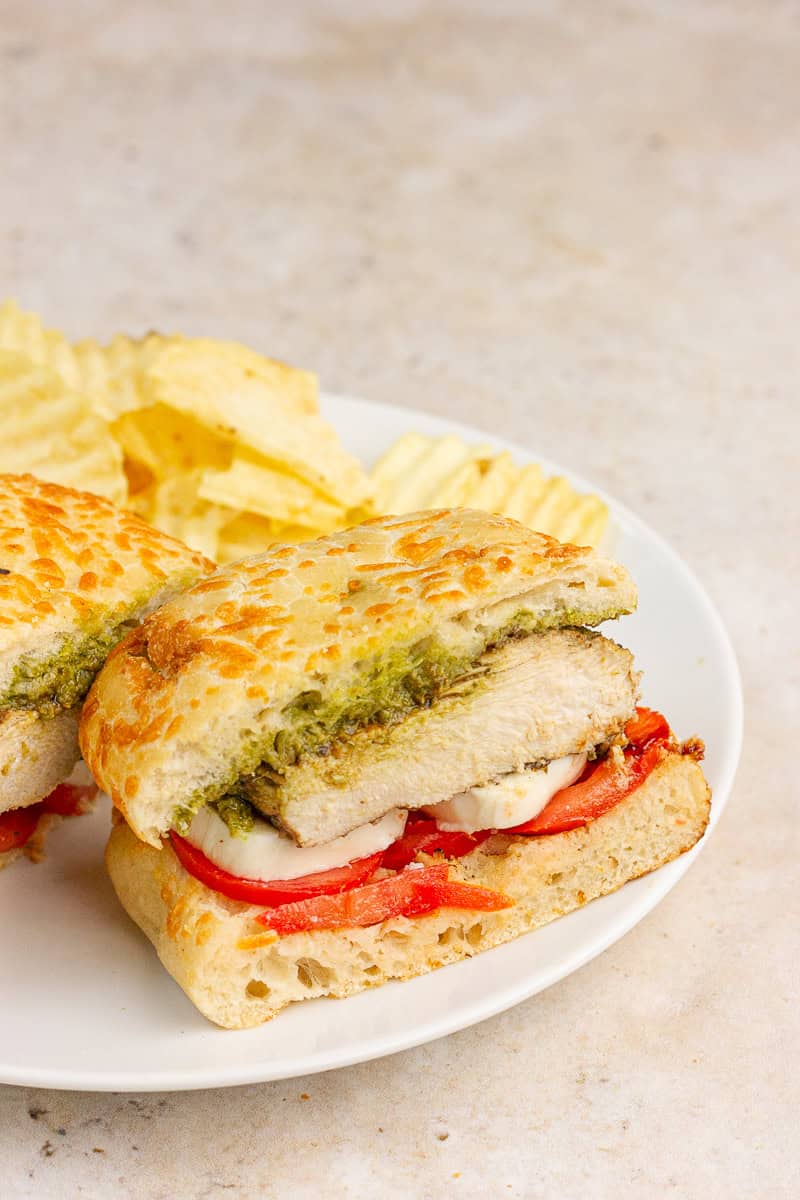 A sliced chicken Caprese sandwich shows layers of pesto, grilled chicken, fresh mozzarella, and tomato on toasted ciabatta.