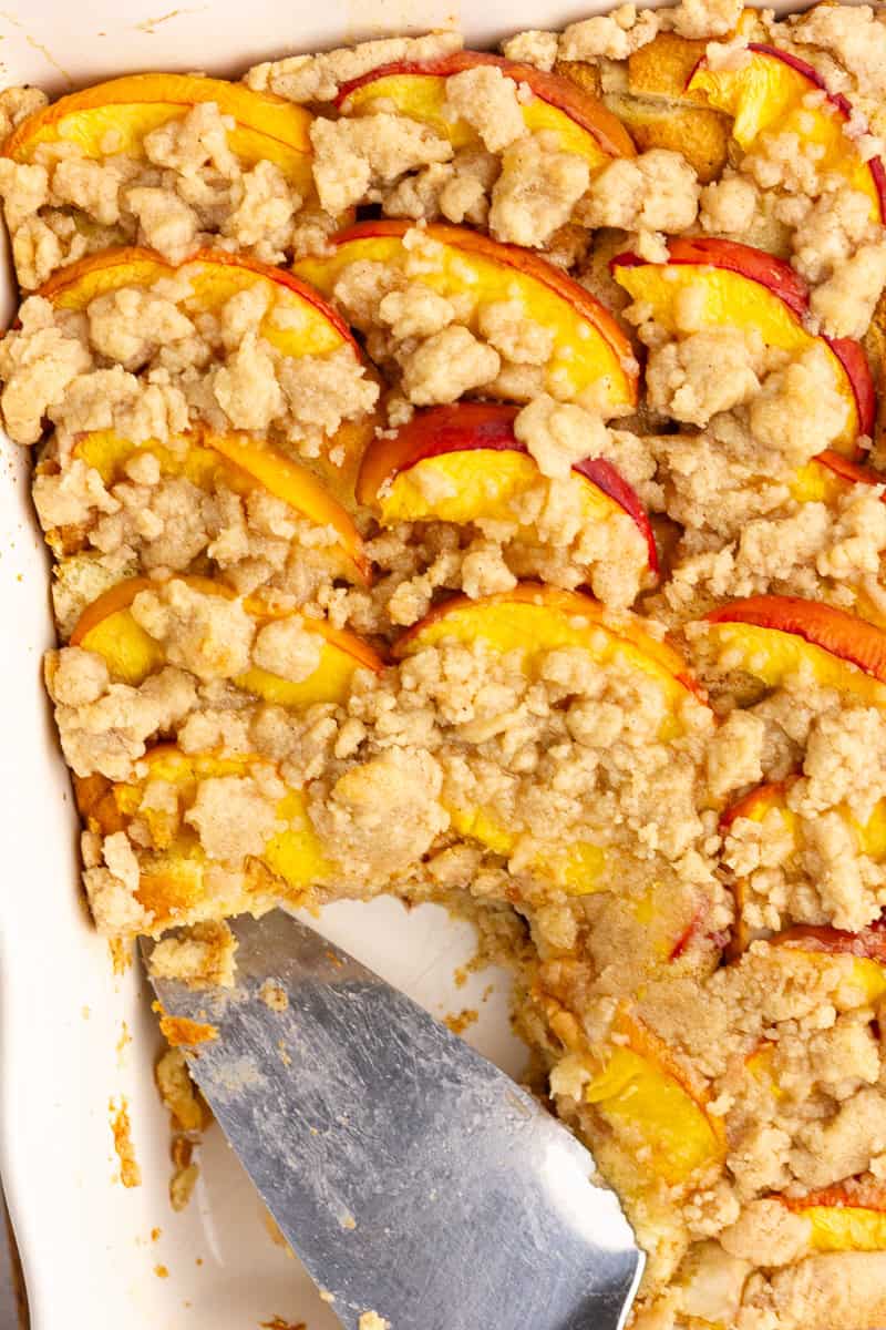 A serving is lifted from the baked peach cobbler French toast casserole, revealing the soft custardy center beneath the crumb topping.
