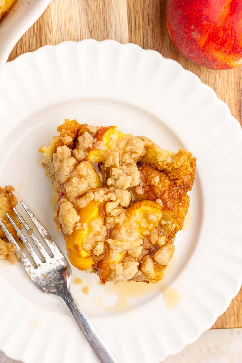 A slice of peach cobbler French toast casserole topped with maple syrup sits on a white plate ready to serve.
