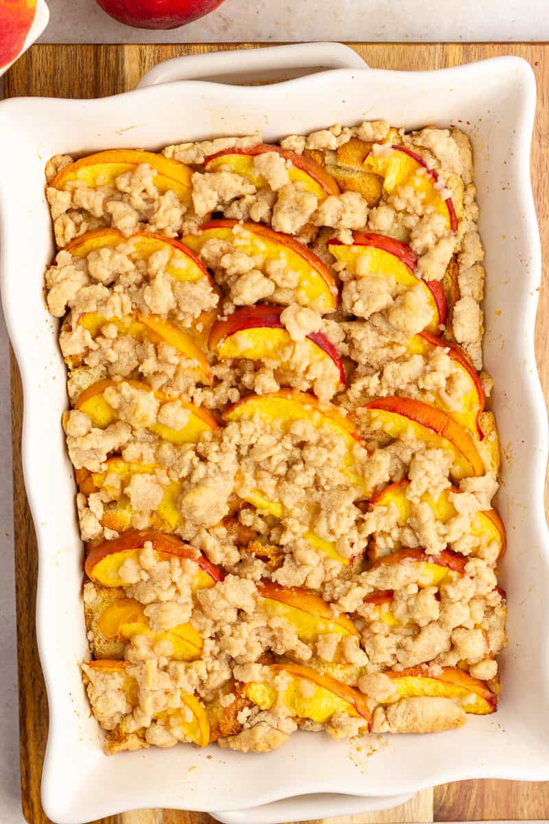Baked peach cobbler French toast casserole is topped with fresh peaches and a golden crumb topping in a baking dish.