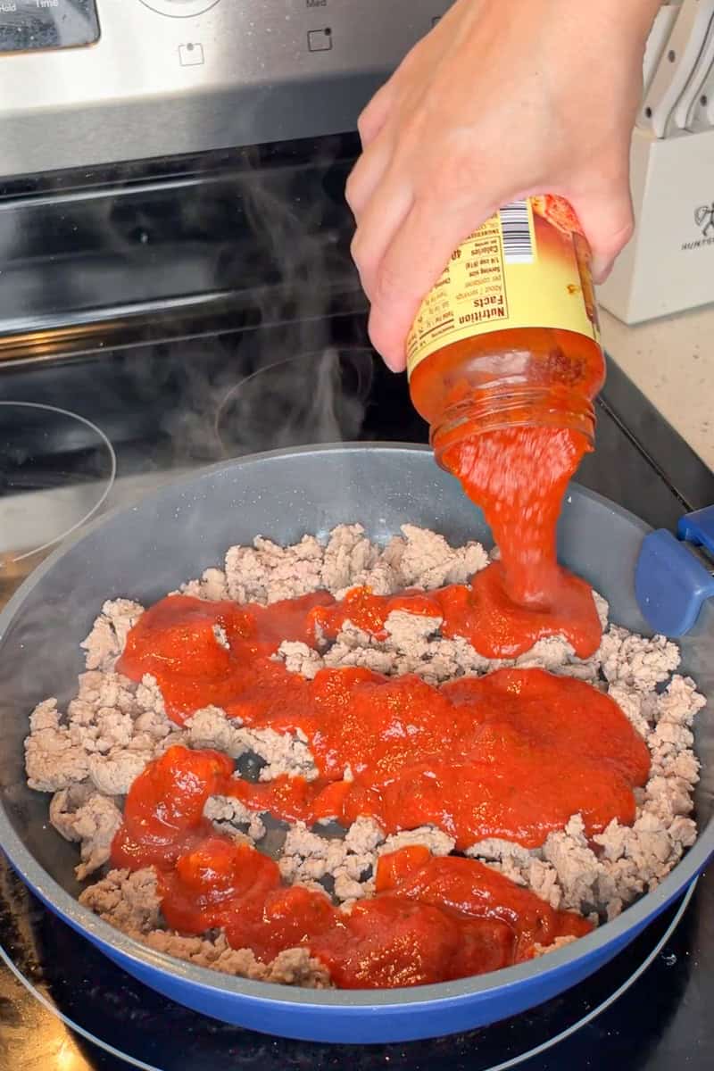 Pizza sauce is poured into a skillet with cooked ground turkey for the casserole filling.