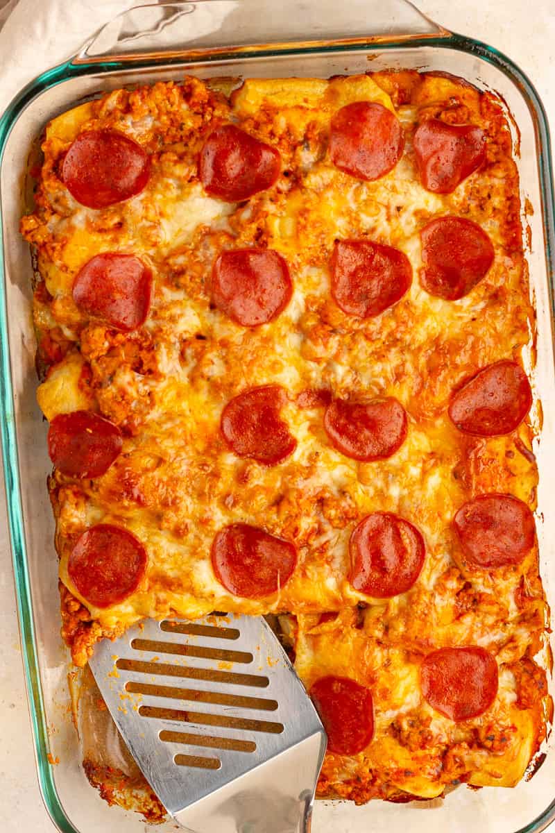 A spatula sits in the baked biscuit pizza casserole topped with melted cheese and turkey pepperoni.