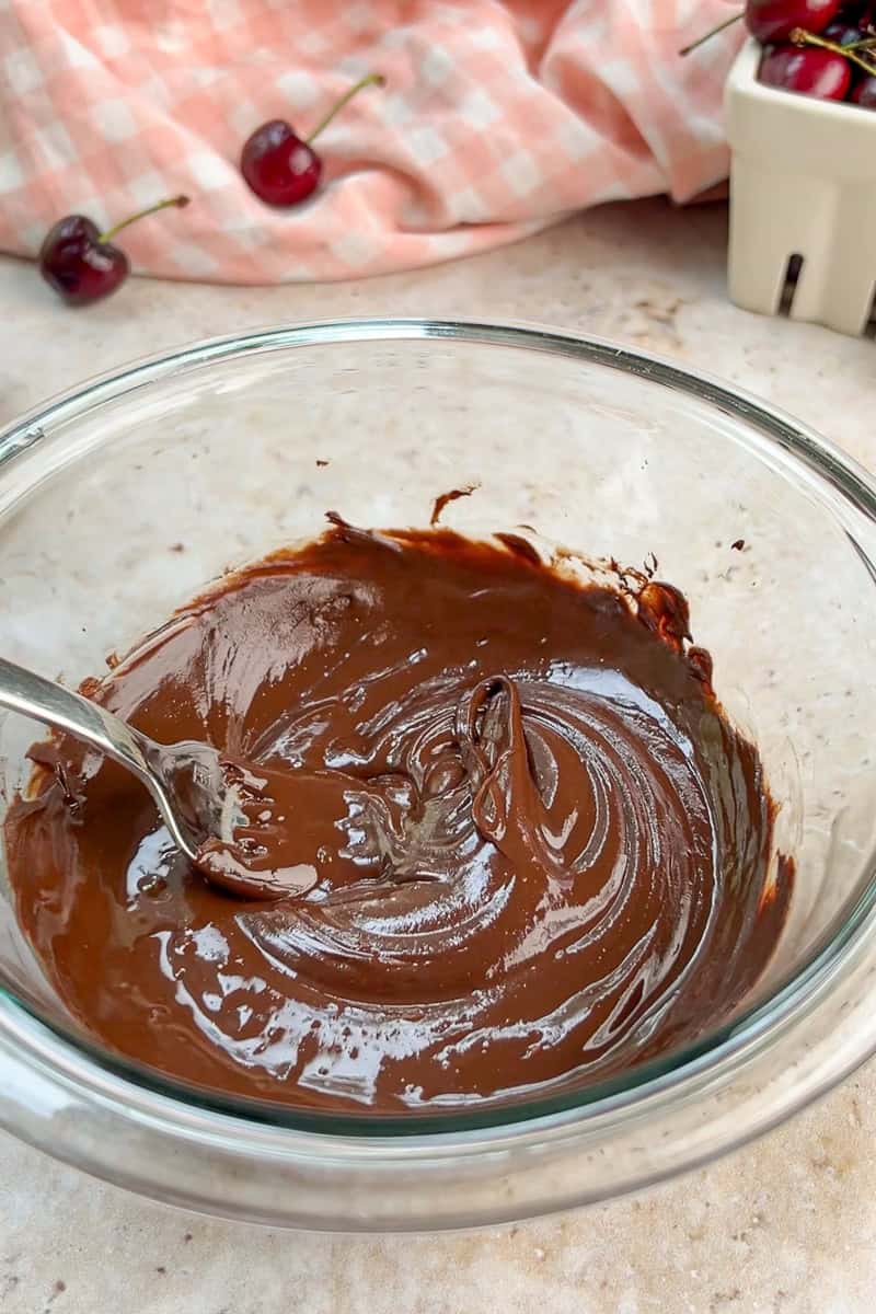 Melted dark chocolate is stirred in a glass bowl for coating yogurt bites.