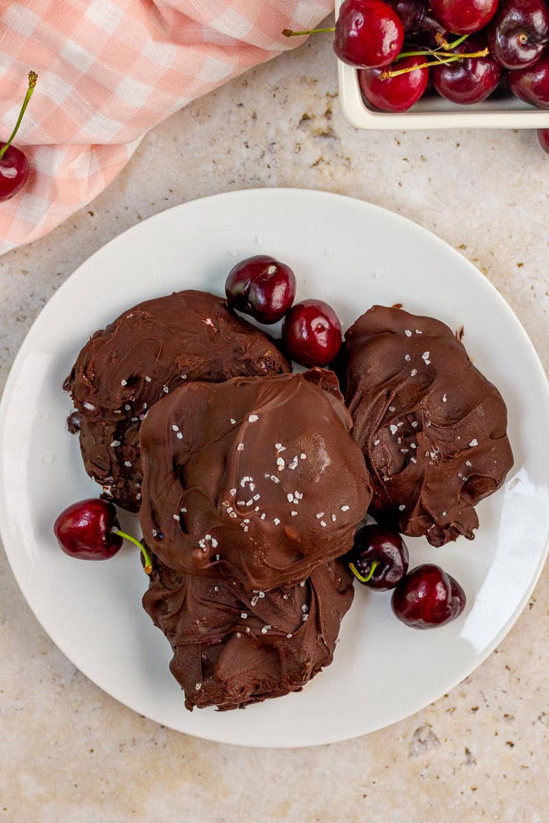 Chocolate covered frozen yogurt bites are served on a white plate with fresh cherries.