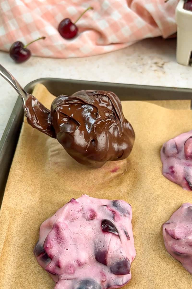 A frozen cherry yogurt bite dipped in melted chocolate is being placed on parchment paper.