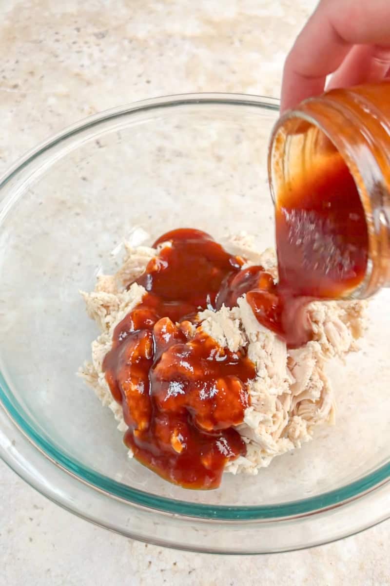 BBQ sauce is poured over shredded cooked chicken in a glass mixing bowl.