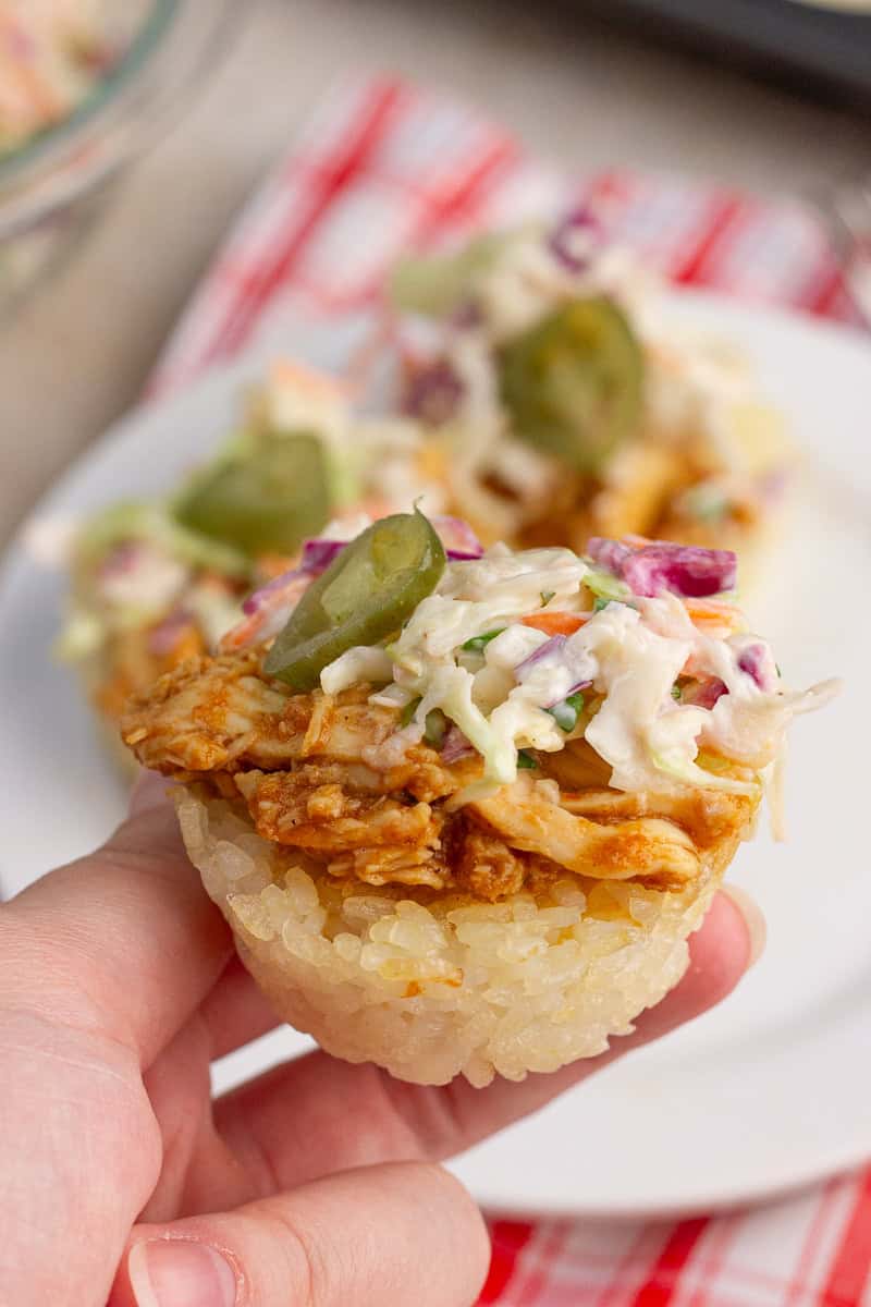 Hand holds a BBQ chicken crispy rice cup topped with ranch coleslaw and pickled jalapeños.