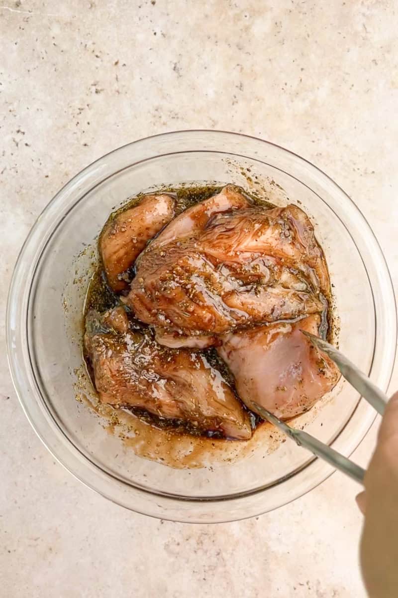 Chicken breasts are tossed in a glass bowl with balsamic marinade using metal tongs.