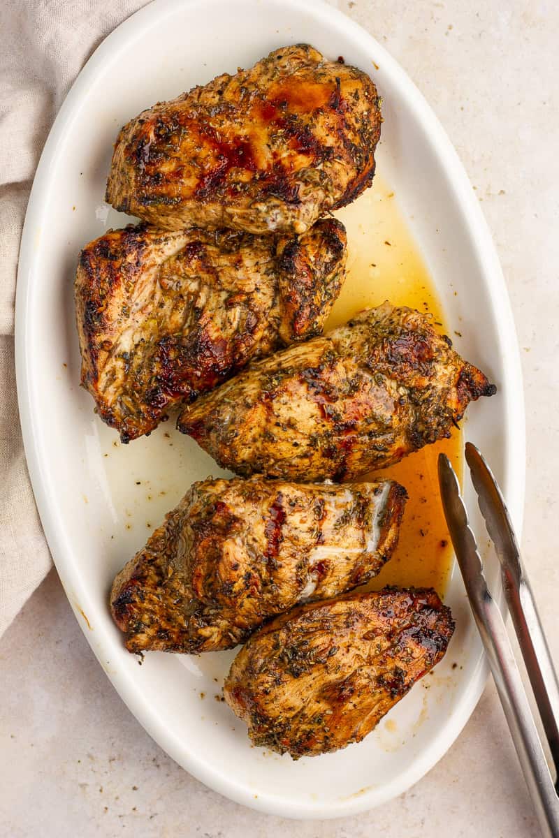 Grilled balsamic chicken breasts are served on a white platter with pan juices and metal tongs.