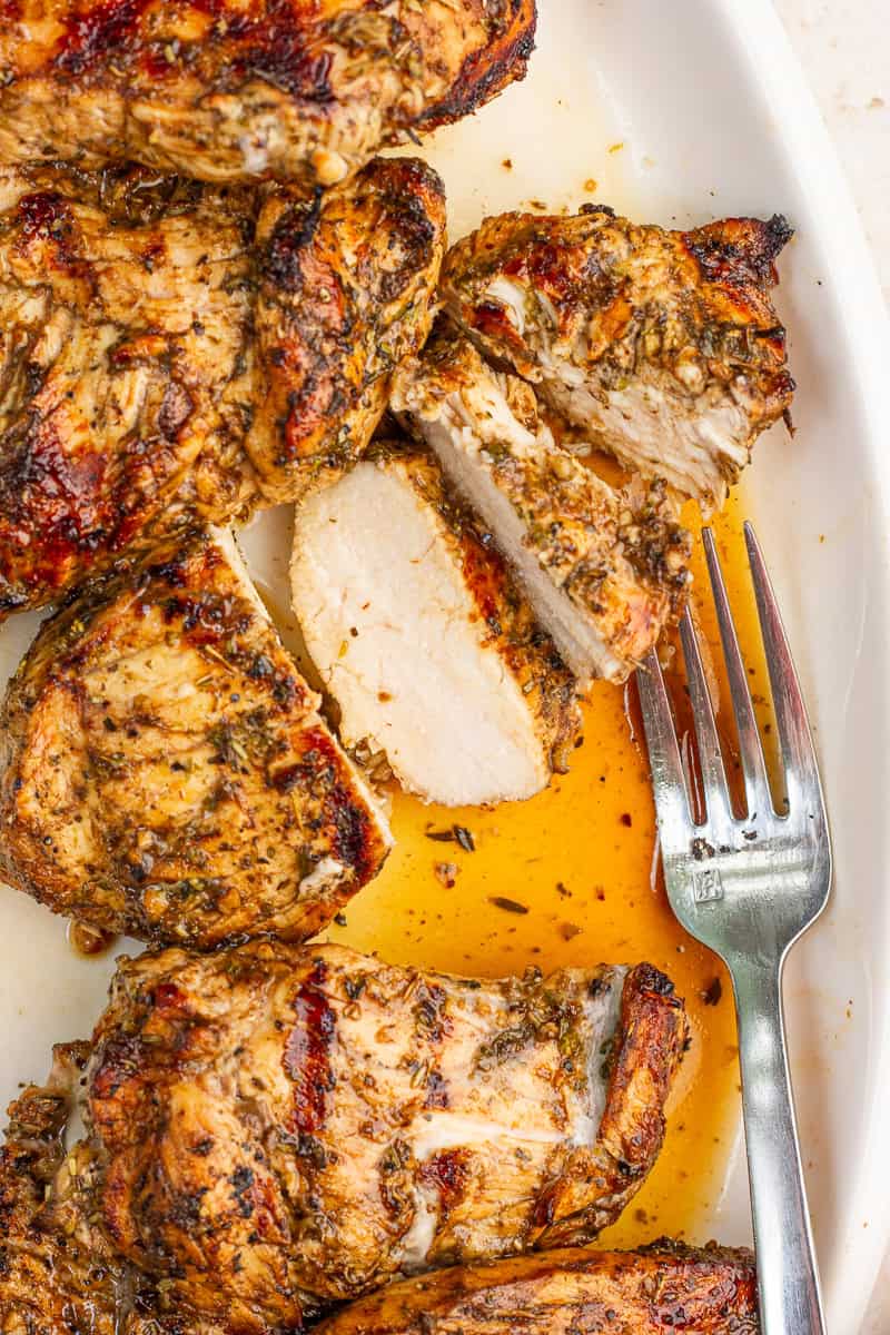 Sliced grilled balsamic chicken breast is shown on a white platter with a fork and flavorful juices.