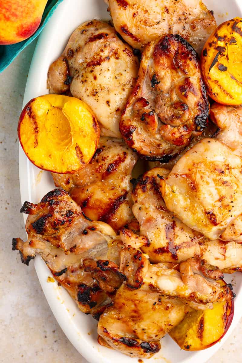 Grilled peach chicken thighs are served on a white platter with charred peach halves.