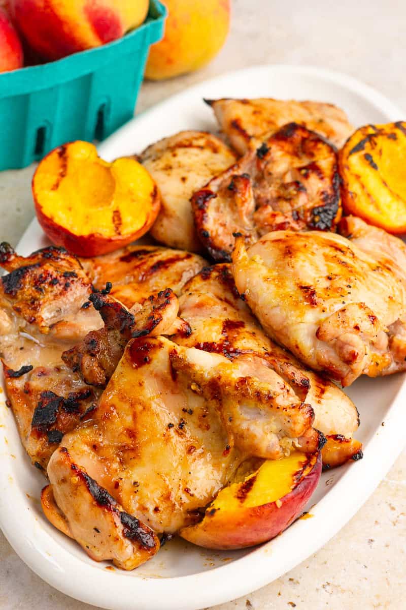 Grilled peach chicken thighs are arranged on a platter with grilled peaches and fresh peaches in the background.