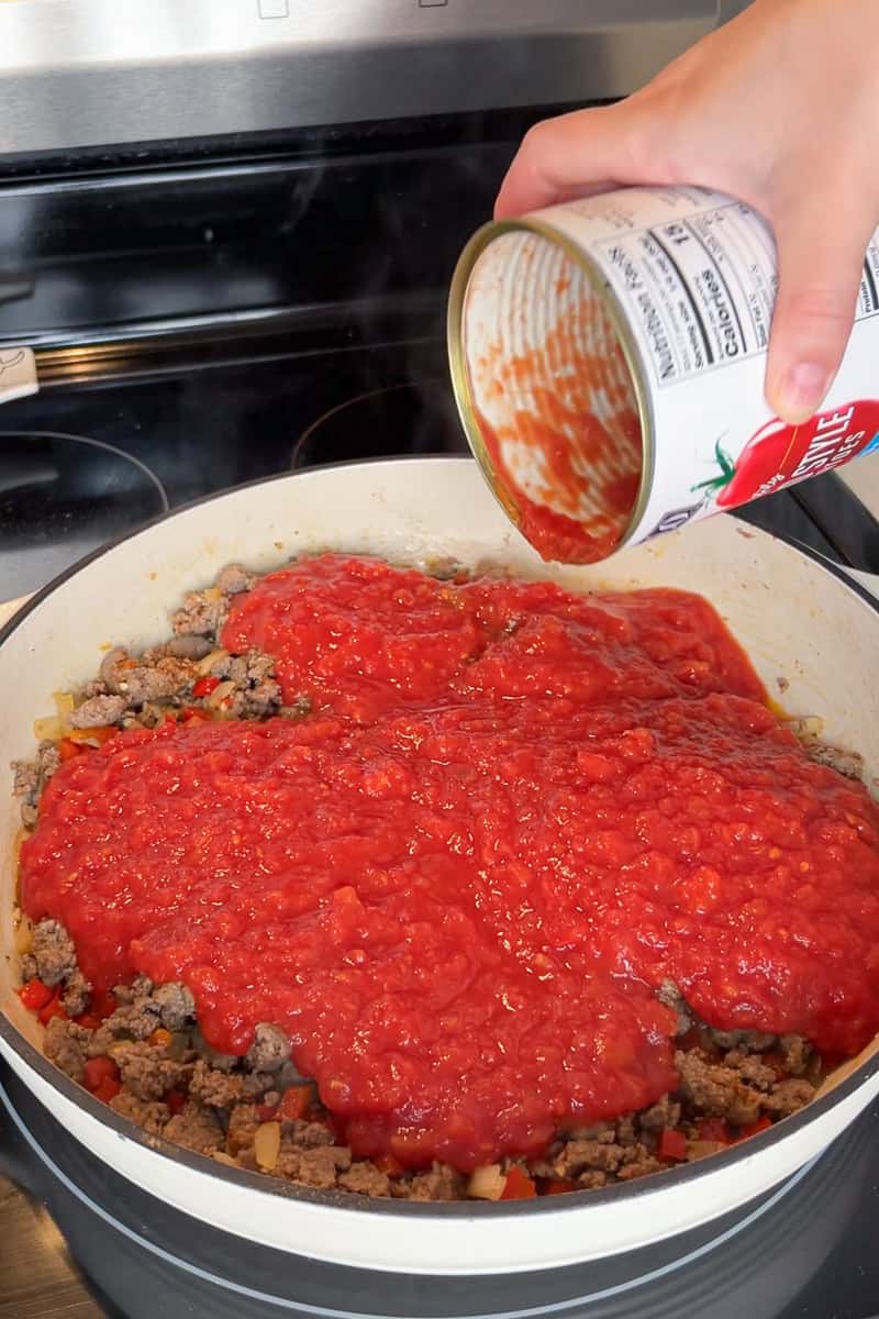 Crushed tomatoes are poured into a skillet with cooked turkey sausage, onion, and red bell pepper.