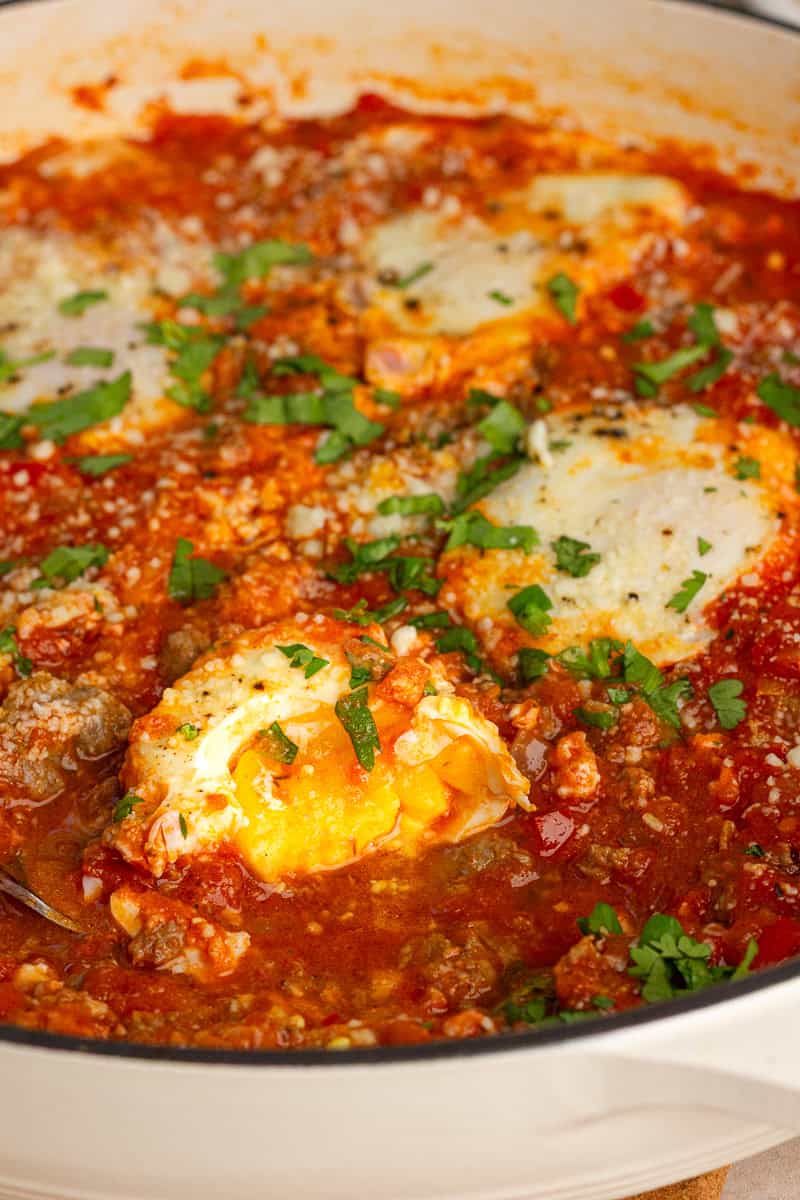 A spoon scoops into high protein shakshuka with eggs, tomato sauce, turkey sausage, feta, and parsley.