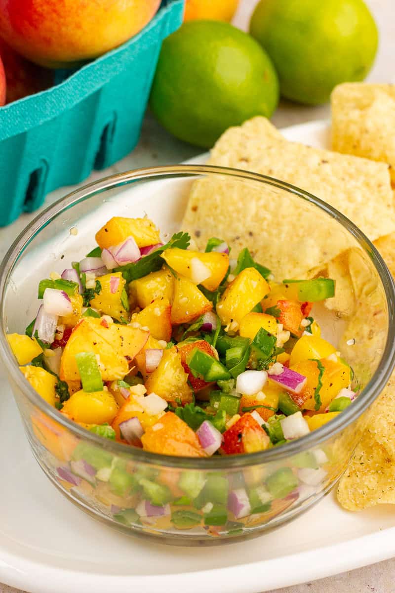 A bowl of jalapeno peach salsa is served with tortilla chips, fresh peaches, and limes.