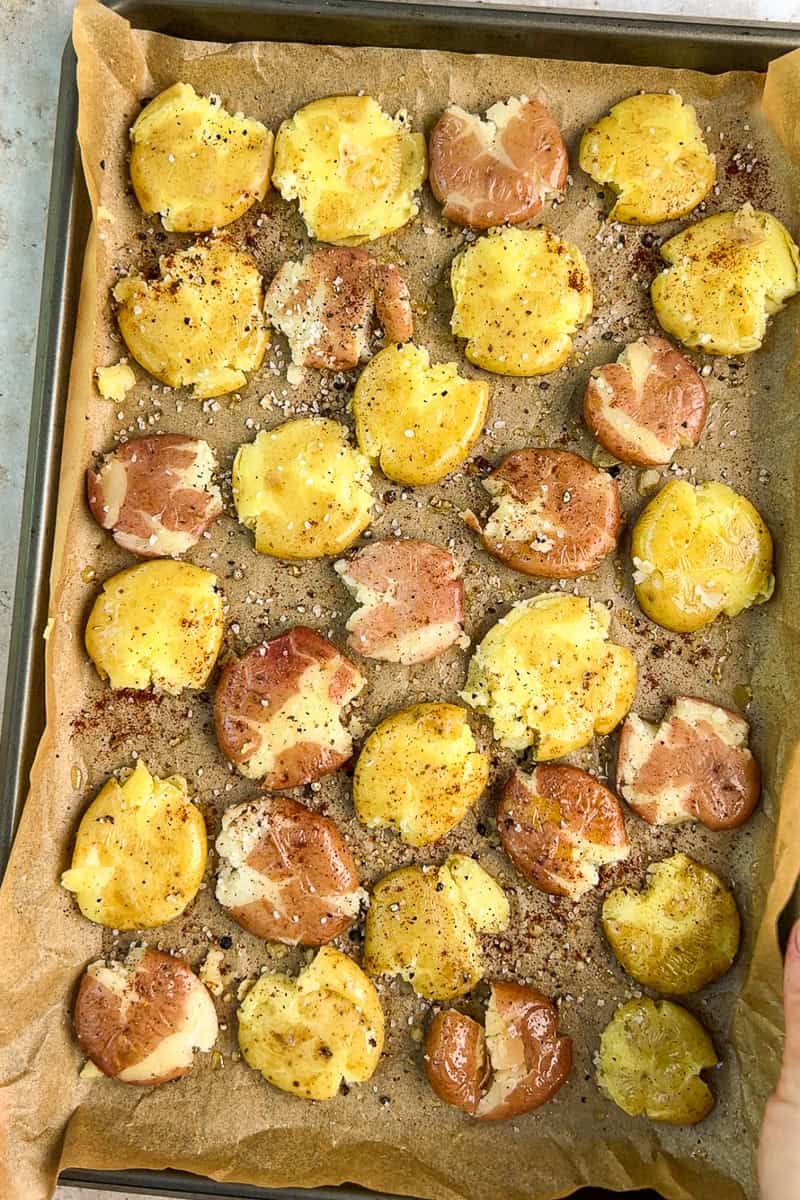 Smashed petite potatoes are seasoned on a parchment-lined baking sheet before roasting.