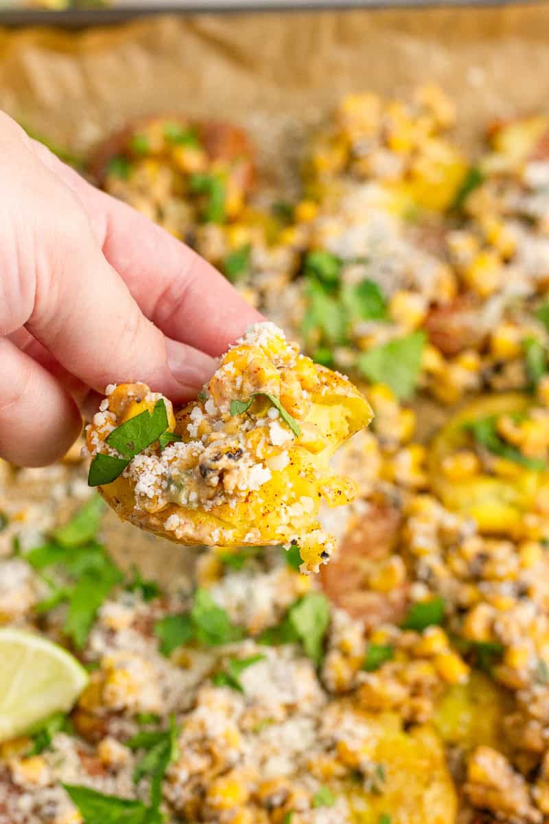 A hand holds a crispy Mexican smashed potato topped with fire roasted corn, cotija cheese, and cilantro.