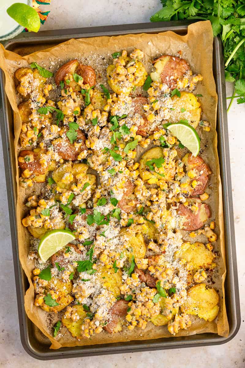 Mexican street corn smashed potatoes are served on a parchment-lined sheet pan with lime wedges and cilantro.