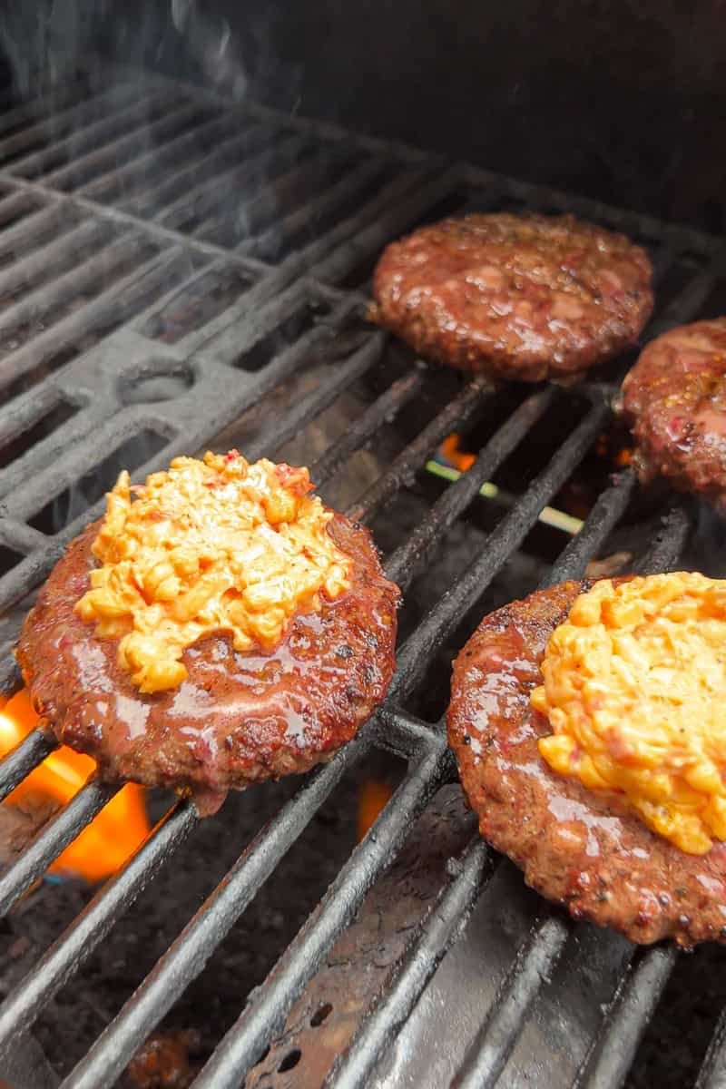 Grilled burger patties topped with pimento cheese cook over an outdoor grill.