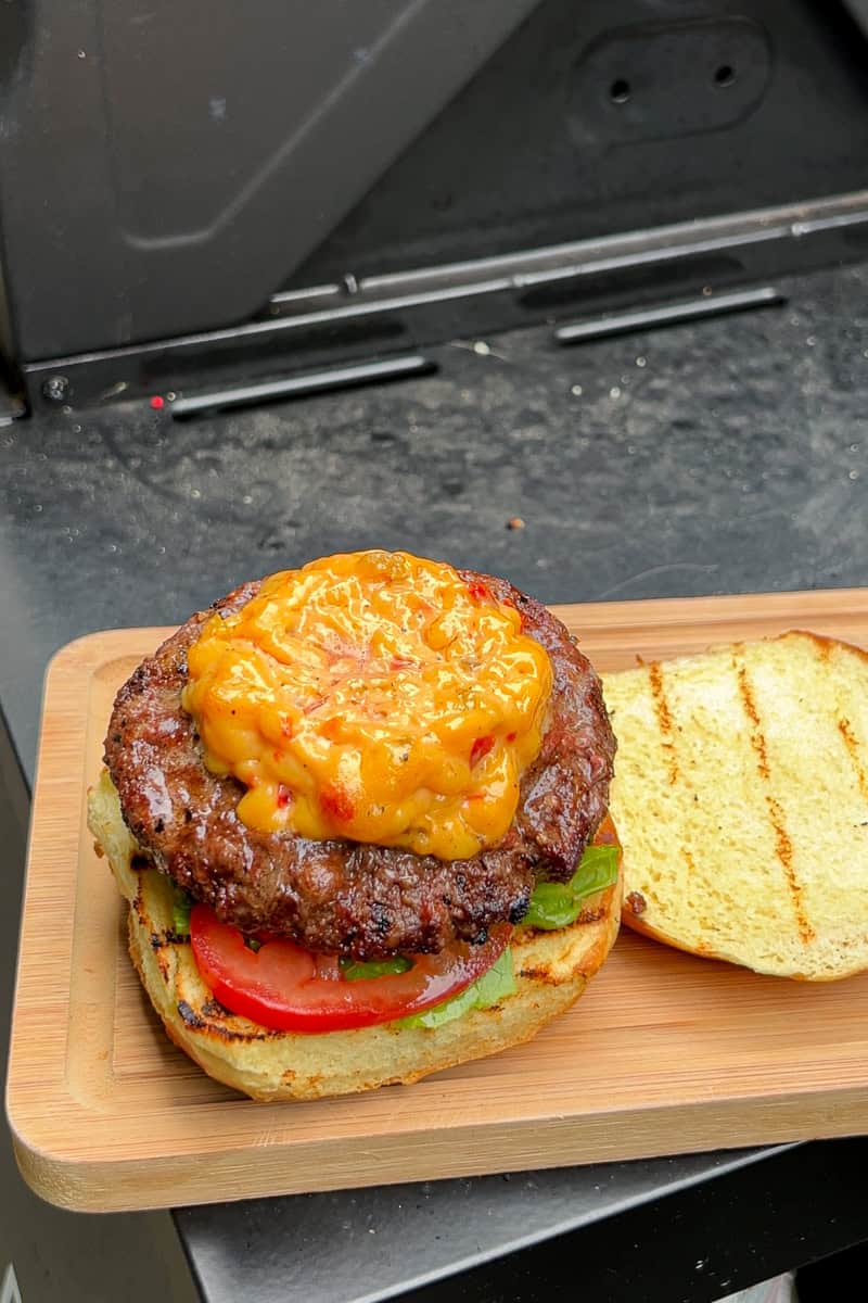 A grilled pimento cheese burger sits on a toasted bun with lettuce and tomato on a wooden board by the grill.