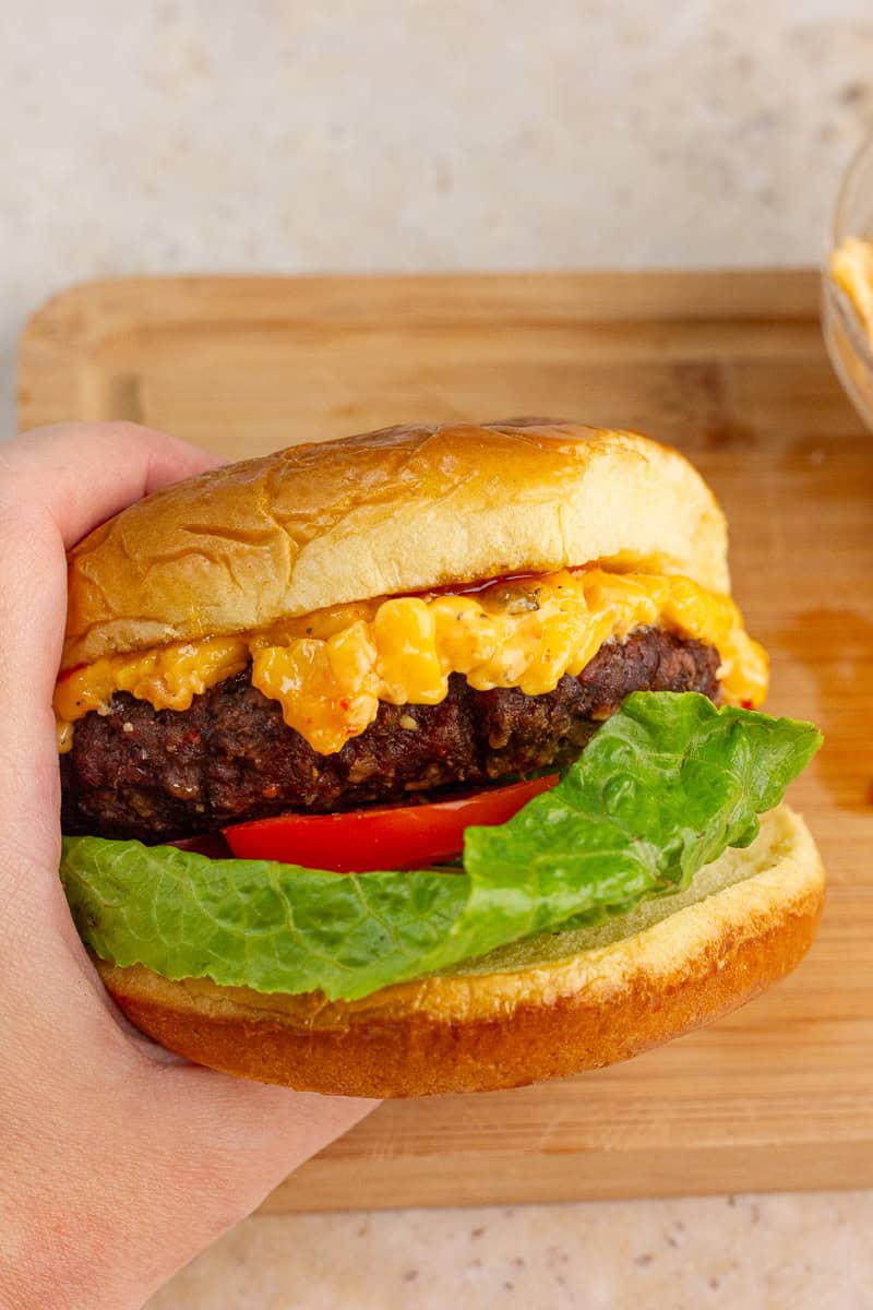A hand holds a pimento cheese burger layered with lettuce, tomato, and melted pimento cheese.