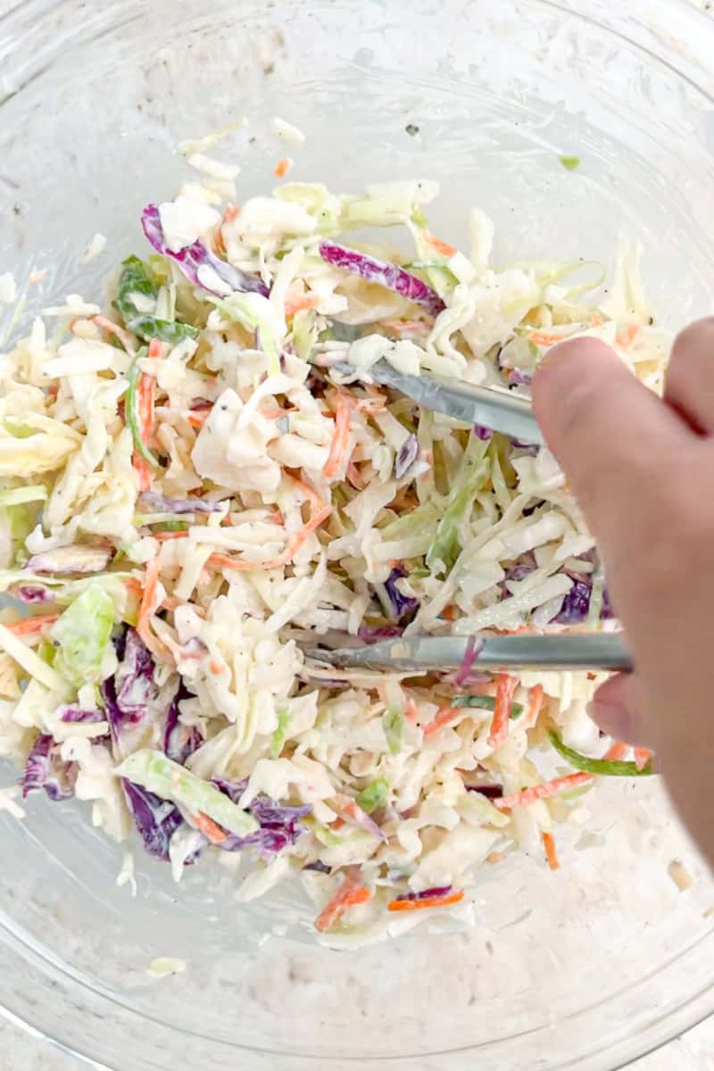 Ranch coleslaw is tossed with tongs in a large glass bowl.